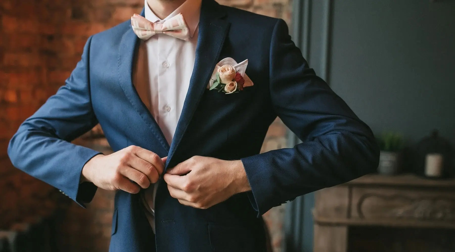 A Caucasian man buttons his navy coat and wears a pink bow tie in front of a brick wall and fireplace.