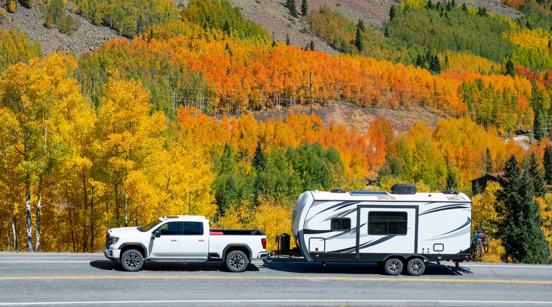 A while pickup truck hauling a large white camper with two bikes on the back, next to a mountain with fall trees.