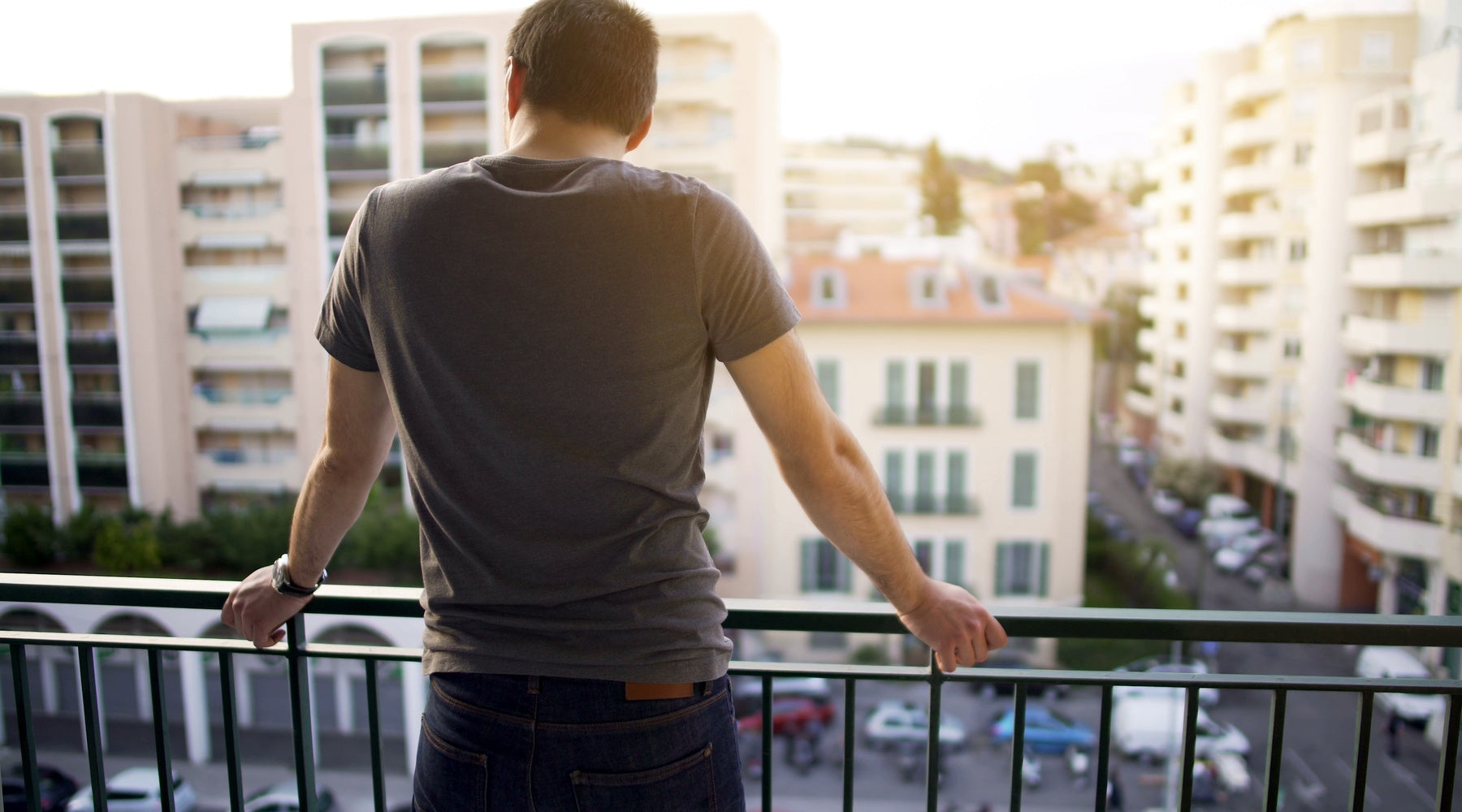 A man grips a balcony railing and looks over apartment buildings from an upper-floor apartment on a sunny day.