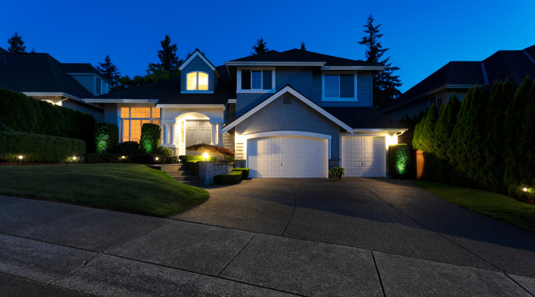 A large suburban home at night with glowing windows, garage lights, a manicured lawn, and a curved driveway.