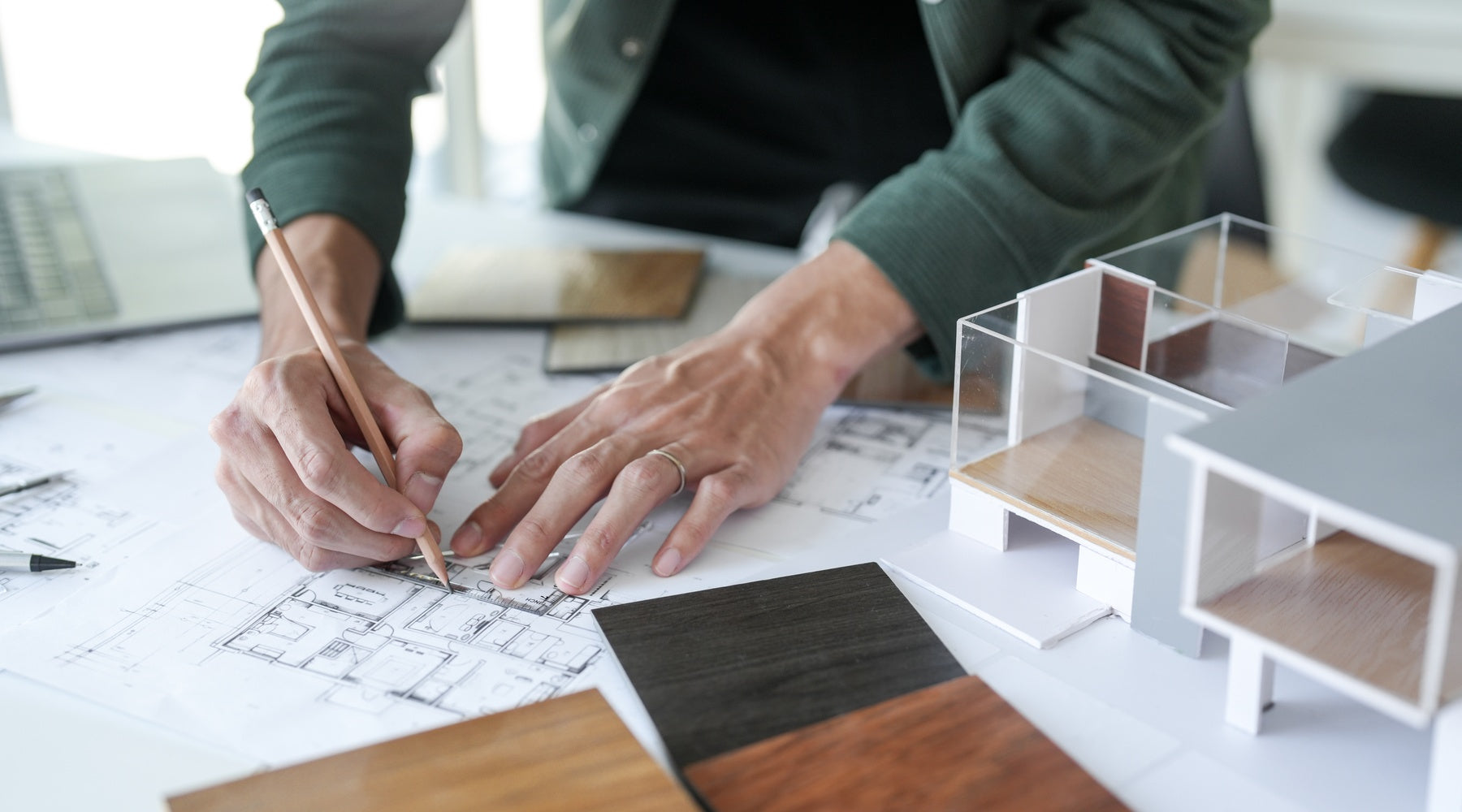 A man reviewing interior design plans on a blueprint with material samples and tools spread across a desk surface.