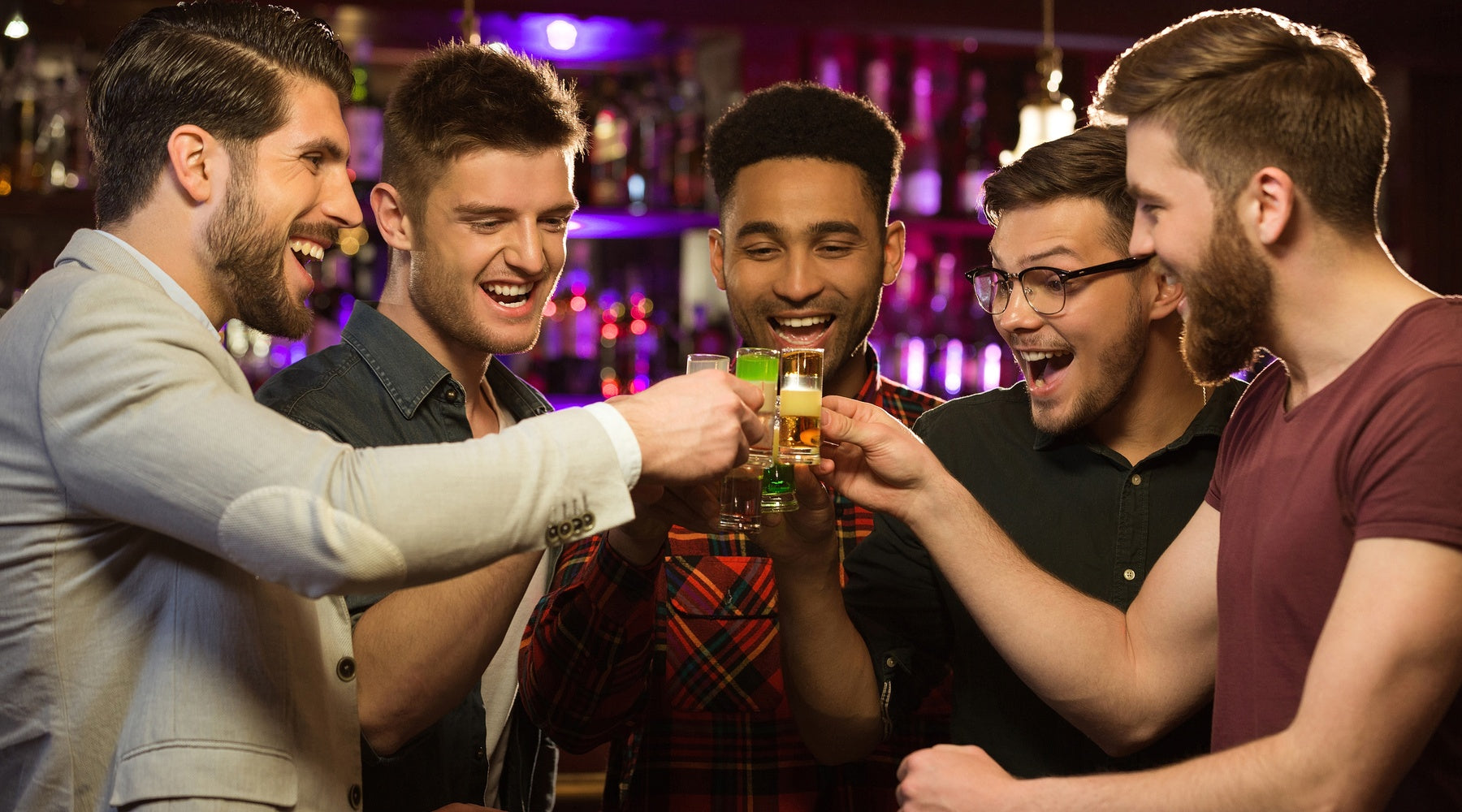 Several men smile while clinking their drinks together around a table inside a casual bar or restaurant.