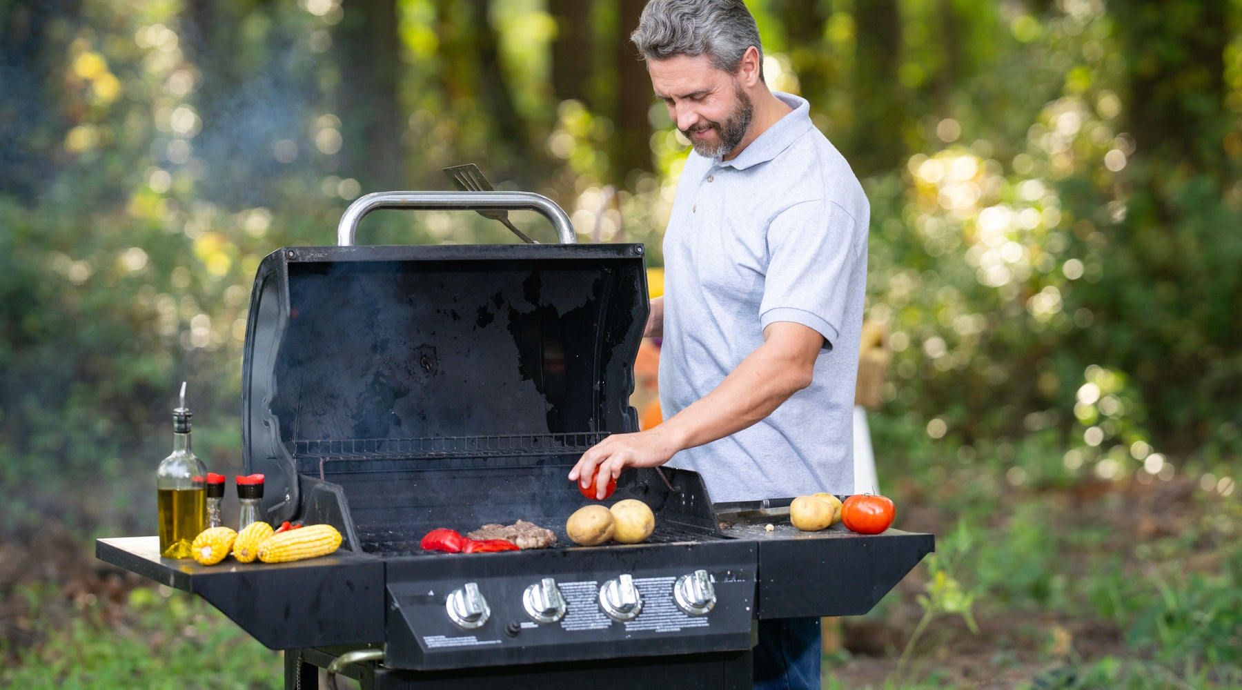 A smiling middle-aged man grills meat and vegetables on a large black gas grill in a wooded outdoor setting.