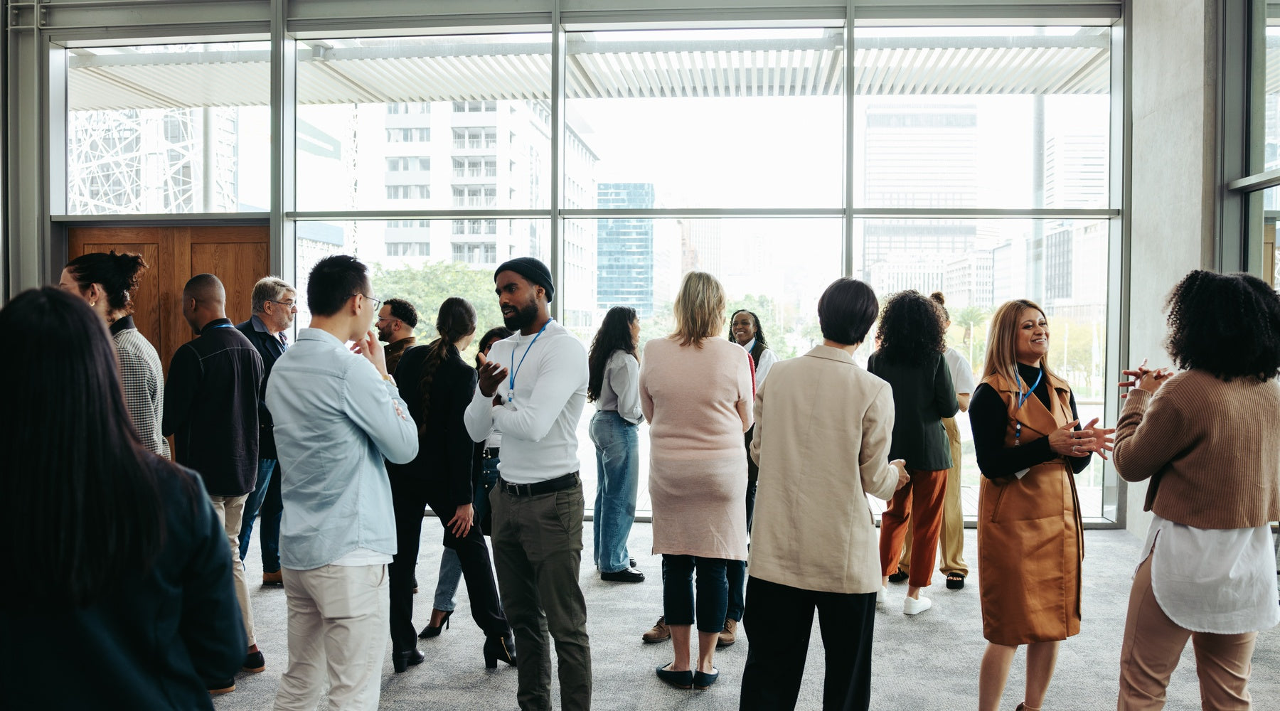 A crowd of people talking inside a large event venue during the daytime. Large glass windows line the walls.
