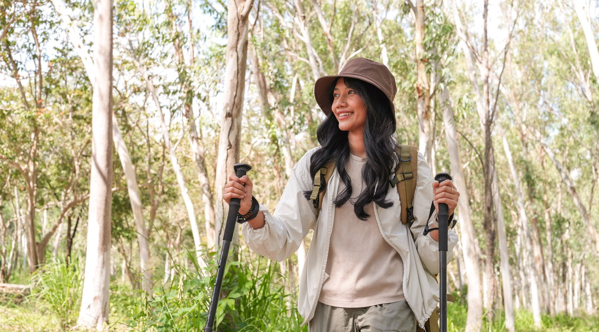 A hiker wearing a brown hat and carrying two thin, black walking sticks in a large forest on a sunny day.