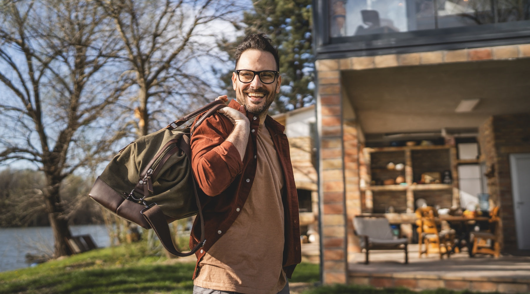 A man with glasses smiles while carrying a travel bag over his shoulder in front of a brick house and bare trees.