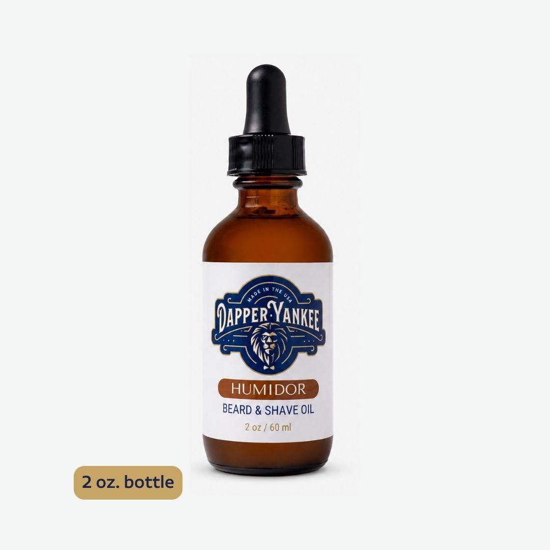 An amber glass dropper bottle of Dapper Yankee Humidor Beard & Shave Oil on a light gray background, with a gold label sticker reading '2 oz. bottle'.