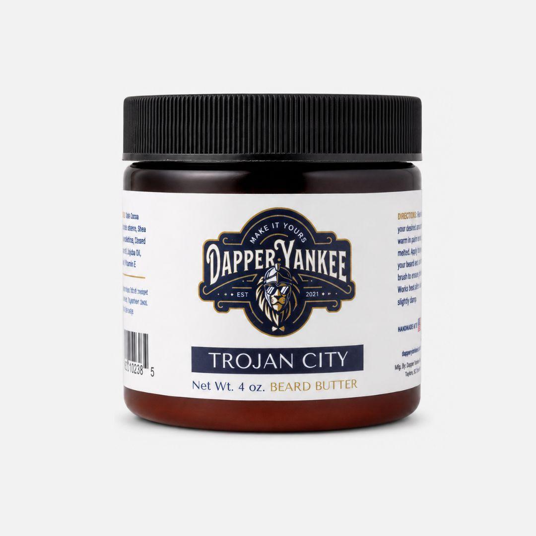 A 4 oz jar of Dapper Yankee Trojan City Beard Butter on a light gray background. The product features a dark amber jar, a black ribbed screw cap, and a white label with navy and gold branding.